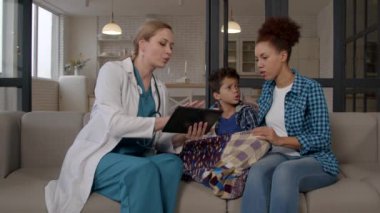 Üniformalı kadın doktor dijital tablet kullanarak tedaviyi açıklıyor. Hasta okul çağındaki Afrikalı Amerikalı çocuk ve endişeli anneye tıbbi prosedürleri gösteriyor..