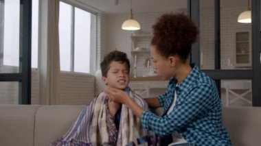 Hasta okul çağında, battaniyeye sarılı Afrikalı Amerikalı bir çocuk. Kanepede oturup, grip ve öksürükten muzdarip ve evdeki boğazı ağrıyan siyah anneye şikayetçi..