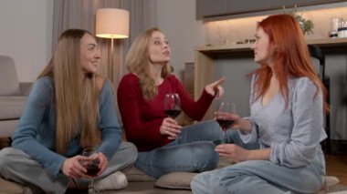 Çekici, zarif, orta yaşlı kadınlar yerde oturuyor, kırmızı şarap içiyor, arkadaşça iletişim kuruyor ve ev ortamında keyif çatıyor..