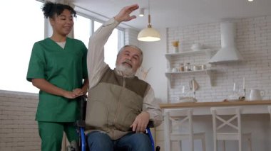 Tekerlekli sandalyedeki yaşlı gri saçlı adam, rehabilitasyon merkezinde rehabilitasyon sırasında siyahi kadın bakıcıdan destek alarak egzersiz yapıyor ve kol kaslarını güçlendiriyor..