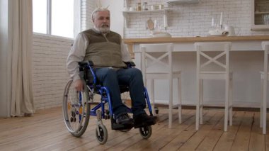 Yaşla ilgili hareket bozukluğu olan yaşlı gri saçlı erkek tekerlekli sandalye kullanıcısı, ev odasında emekliliğinin keyfini çıkarırken kendine güvenini ve pozitifliğini dile getiriyor..