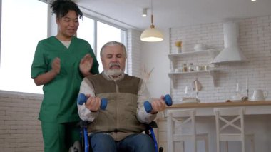 Kararlı yaşlı gri saçlı erkek tekerlekli sandalye kullanımı, yaşla ilgili hareket yetersizliği, güzel siyahi kadın bakıcı, halterli bisepslerin kıvrılması, bakım evinde kol kaslarının güçlendirilmesi