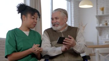 Üst düzey gri saçlı erkek tekerlekli sandalye kullanıcısı ve üniformalı mutlu siyahi kadın bakıcı sosyal medya içeriğini bakımevinde cep telefonuyla tarıyor ve tartışıyor..