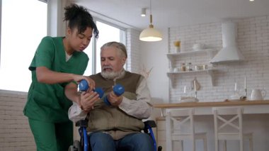 Tekerlekli sandalyedeki yaşlı adam yaşla ilgili hareket engelli Afrikalı Amerikalı kadın bakıcı, halterli egzersiz yapıyor, bakım evindeki kol kaslarını güçlendiriyor..