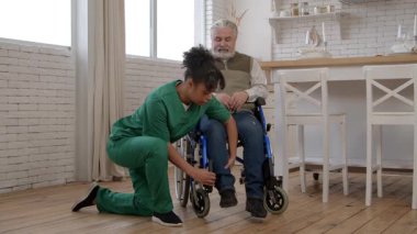 Çekici siyahi kadın bakıcı üniformalı yaşlı, gri saçlı, hareket bozukluğu olan bir adamla ilgileniyor, uzun süreli bakım merkezlerinde hemşirelik yaparken tekerlekli sandalyeye ayak basmaya yardım ediyor.