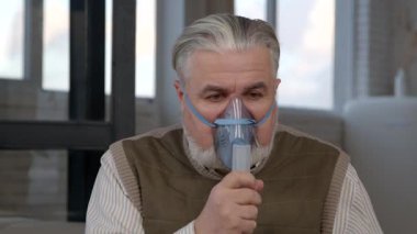 Astım ve solunum hastalıklarından muzdarip oksijen maskeli yaşlı, gri saçlı hasta bir adamın portresi, bakım evinde jet nebulizatörüyle solunum terapisi yapıyor..