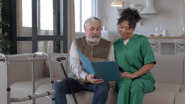 Afro-Amerikan kadın bakıcıya eski günlerden bahsederken evdeki kanepede keyfine varan, eski günlerden bahseden, klasik fotoğraflı, yaşlı, çekici, gri saçlı bir adam..