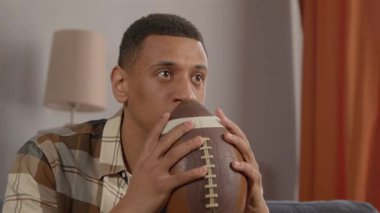 Mutlu siyah erkek Amerikan futbol fanatiğinin portresi TV 'de futbol maçı izliyor, favori takımı destekliyor ve galibiyeti jestlerle kutluyor, içeride heyecan ve neşeyi ifade ediyor.