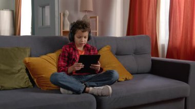 Kablosuz kulaklıklı Afro-Amerikalı genç çocuk kanepede bağdaş kurarak oturuyor, tablet bilgisayarında oyun oynuyor ve evdeki takım arkadaşlarıyla iletişim kuruyor..