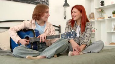 Akustik gitarlı neşeli genç kız ve mutlu kızıl saçlı orta yaşlı anne yatakta bacak bacak üstüne atmış, arkadaşça konuşuyor ve eğlenirken evde eğleniyor..