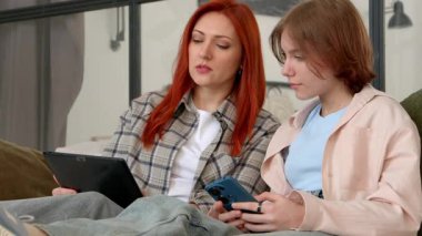 Tabletli güzel, kızıl saçlı olgun anne portresi ve cep telefonlu sevimli kız çocuğu internette sosyal medya içeriğini tartışırken evde keyif çatıyor..