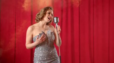 Şık elbiseli, mikrofonlu çekici bir bayan şarkıcı. Sahnede canlı performans sergilerken neşeli bir şarkı söylüyor..