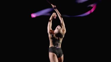 Şık taytlı, parlak kurdeleli, becerikli, ritmik jimnastikçinin arkadan görünüşü geriye doğru eğiliyor. Kılıç ustasının sarmal unsurlarını kafasının üstünde sergiliyor. Siyah arka planda yılanlar var.