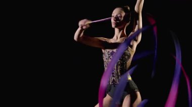 Şık taytlı, zarif ritmik jimnastikçinin portresi. Kurdeleli, siyah arkaplanda spiral elementler sergiliyor..
