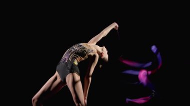 Tarz sahibi, zarif, esnek, şık bir bayan sporcunun kurdeleli arka planda ritmik jimnastik yılanları uygulamasının yan görüntüsü.