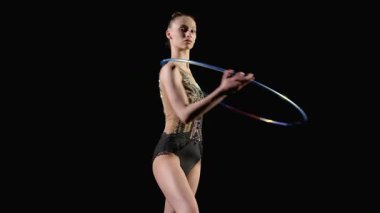 Zarif, formalı, şık bir kadın sporcunun portresi ritmik jimnastik elementler sergiliyor. Boynunun etrafında dönen parlak bir halka. Tek eliyle siyah arka planda dönüyor..