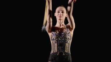 Zarif kadın sporcunun zarif bir portresi şık bir tayt giymiş ritmik jimnastik elementler uyguluyor, her iki elin arasında siyah arka planda sopalarla değirmenleri icra ediyor.