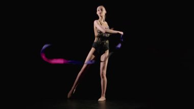 Zarif, çekici ritmik jimnastikçi elinde parlak bir kurdele, etrafında dönüyor ve siyah arka plandaki performans sırasında vücudun etrafında tam bir kumaş çemberi oluşturuyor.