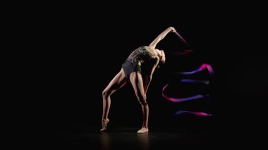 Zarif taytlı, esnek bayan sporcu. Geriye doğru eğiliyor. Siyah arka planda kurdeleli ritmik jimnastik yılanları kullanıyor.