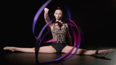 Esnek profesyonel çekici ritmik jimnastikçi yan yana oturmuş, siyah arka planda parlak kurdeleli spiraller sergiliyor..