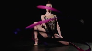 Şık giyimli, şık bir jimnastikçinin portresi düşük hamlelerde oturup siyah arkaplan üzerinde spiral elementlerle desenli kurdeleli yılanlar üzerinde pratik yapıyor..