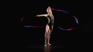 Zarif profesyonel çekici ritmik jimnastikçi parlak kurdeleli zarif bir mayo tutuyor, onu döndürüyor ve siyah arka planda performans sırasında vücudun etrafında bir kumaş çemberi oluşturuyor.