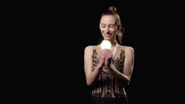 Havalı, zarif taytlı genç bayan sporcu ritmik jimnastik yarışmasını kazandıktan sonra elinde altın madalyayla siyah arka planda mutluluk ve neşe ifade ediyor..