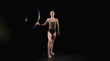 Son derece yetenekli, zarif, ritmik jimnastikçi şık taytlarıyla jimnastik yapıyor, iki eliyle dönen ve hokkabazlık yapan, siyah arka planda arkaya pas veren.