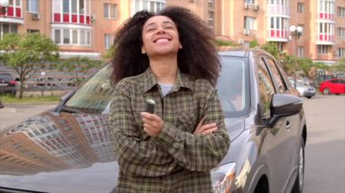 Çekici, Afro-Amerikan kadın sürücü, araba kaputuna yaslanıyor, yeni aracın anahtarsız giriş sabitleyicisini tutuyor, araba galerisinde alışveriş yaptıktan sonra heyecan ve neşe ifade ediyor..