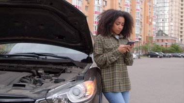 Bozuk aracın yanında duran güzel siyah kadın sürücü, cep telefonuyla internette geziniyor ve şehirde arabayla seyahat ederken yol kenarı yardım servisi arıyor..