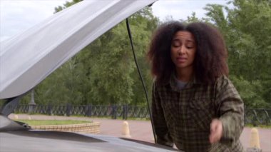 Kafası karışmış Afro-Amerikan kadın sürücü açık araba kaputunun yanında duruyor, bozuk araç motorunu tamir etmeye çalışıyor ve yolculuk sırasında yardım istiyor..