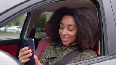Çekici Afrikalı Amerikalı kadın sürücünün şehir caddesinde araba sürüşünün portresi, arkadaşınla internette cep telefonu kullanarak görüntülü görüşme, yolculuk sırasında pozitifliği ve arkadaşlığı ifade etmek..