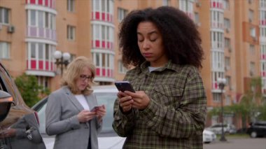 Üzgün Afrikalı Amerikalı kadın portresi hasarlı aracın yanında duruyor, cep telefonuyla internette geziniyor ve arka planda suçlu kadın sürücünün bulunduğu yol kenarı yardım servisi arıyor.