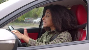 Kızgın, sinirli, Afro-Amerikan kadın sürücü trafik sıkışıklığında sıkışmış, kornaya basıyor, şehirde işlek yolda araç kullanırken el kol hareketi yapıyor ve tartışıyor..