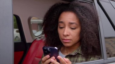 Afro saçlı güzel Afro-Amerikan bir kadın arabanın arka koltuğunda oturuyor, internette kötü haberler okuyor ve kafasını cep telefonuyla destekliyor, hayal kırıklıklarını ve endişelerini dile getiriyor. .