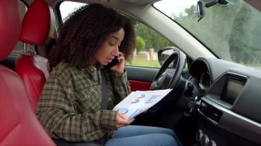 Sürücü koltuğunda oturan, cep telefonuyla konuşan ve yolculuk sırasında hareket halinde olan finansal tabloları inceleyen başarılı, konsantre olmuş siyah kadın yöneticinin portresi..