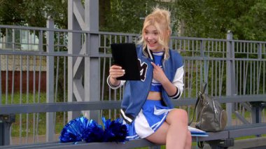 Üniformalı ve beyzbol ceketli popüler sarışın amigo kız dijital tablet kullanarak internetten video görüşmesi yapıyor okuldan sonra tribünde dinlenirken arkadaşlarıyla sohbet ediyor..