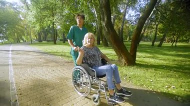 Yaşla ilgili hareket bozukluğu olan güzel, yaşlı kadın tekerlekli sandalye kullanıcısı üniformalı siyahi kadın bakıcı yaz parkında yürürken temiz havanın ve eğlencenin tadını çıkarıyor..