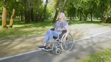 Yaşına bağlı hareket engelli, aktif, çekici, yaşlı bir kadın. Park sokağında tekerlekli sandalyeyle dolaşırken boş zamanların, temiz havanın ve yaz doğasının tadını çıkarıyor. ..