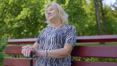 Yaşla ilgili hareket engelli, pozitif çekici yaşlı kadın bankta oturuyor, baston tutuyor, yaz parkında sabah yürüyüşü sırasında boş vaktini ve temiz havayı harcıyor..