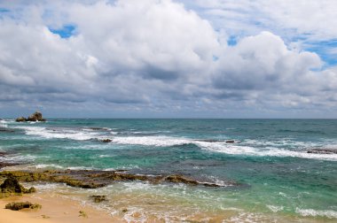 Gök mavisi okyanusu ve mercan kayalıkları olan tropik bir sahil. Kümülüs bulutlarıyla bulutlu bir gün.