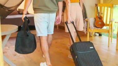 Tilting up rear of young Biracial woman and man carrying luggage, arriving at their tent in glamping on sunny summer day