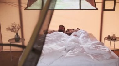 Young dark-haired Biracial woman and bearded husband waking up in bed in glamping, stretching and hugging in morning
