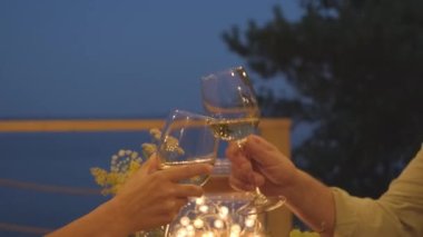 Side view medium of cropped woman and her husband clinking glasses with white wine, sitting at table on terrace by lake at night, couple celebrating anniversary