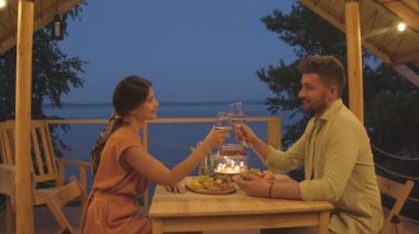 Side view medium of elegant Biracial man clinking glasses with white wine with his beautiful wife, sitting at table on terrace by lake at night, couple having romantic date