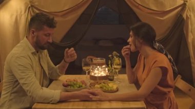 Side view medium of elegant Biracial woman and young husband drinking wine, holding hands, talking and snacking, sitting at table in glamping at night, couple having romantic date