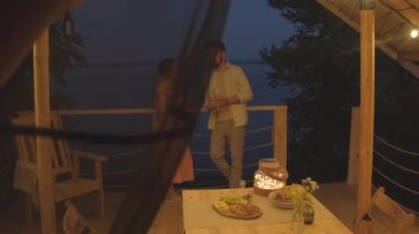 Long shot of beautiful Biracial woman and young husband standing and talking on terrace by lake at night, drinking wine on relationship anniversary on vacation