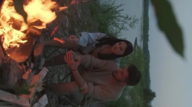 Vertical of young bearded Biracial man sitting with young girlfriend on lakeshore by campfire on summer evening, roasting sausages on skewers, talking and enjoying nature
