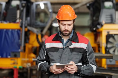 Serious young foreman or technician in safety helmet and uniform scrolling through online technical information in tablet