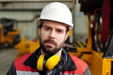 Serious engineer of industrial plant in safety helmet looking at camera against part of huge machine inside large workshop